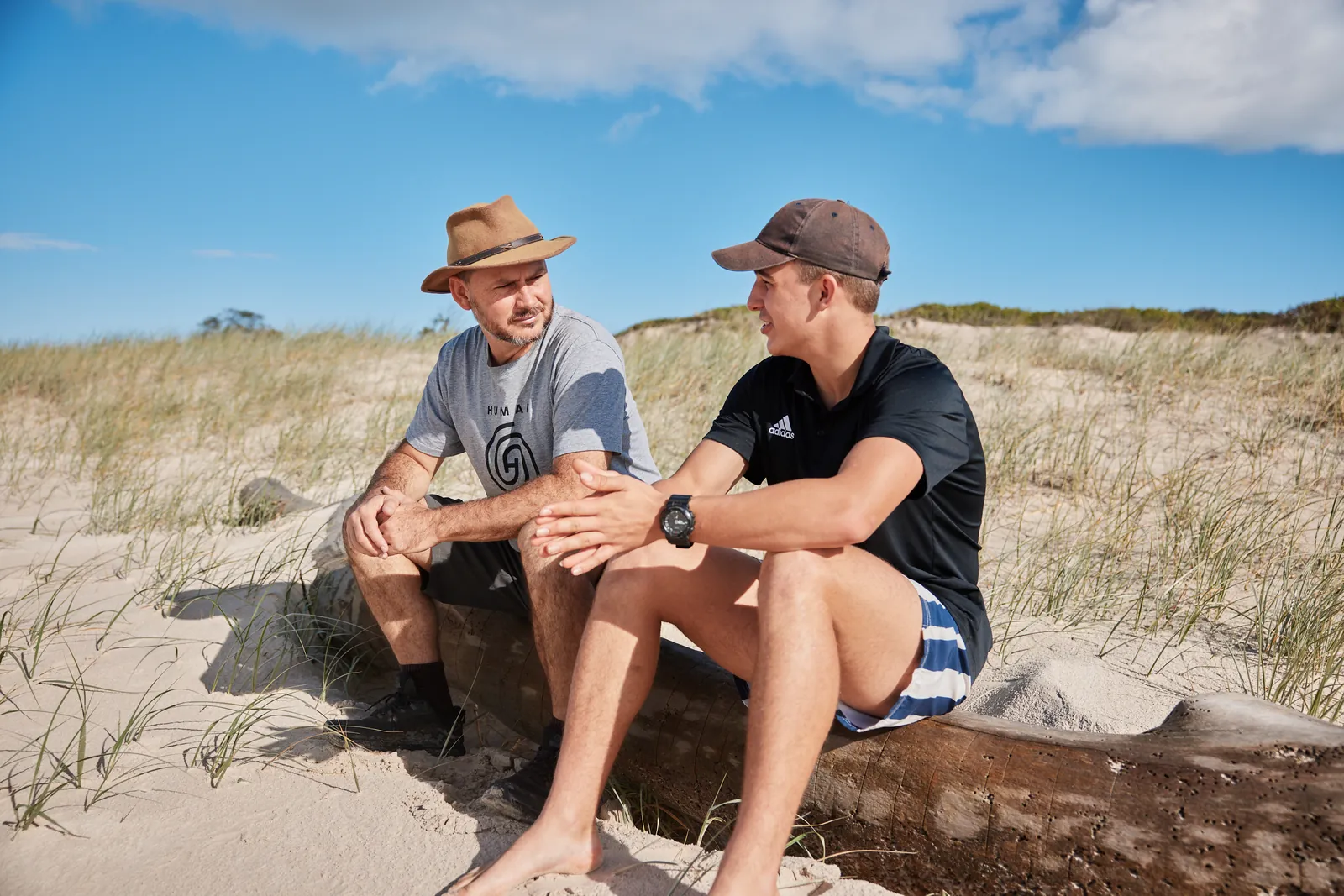 A mentor and a young man sitting side by side on a fallen tree in the dunes, talking
